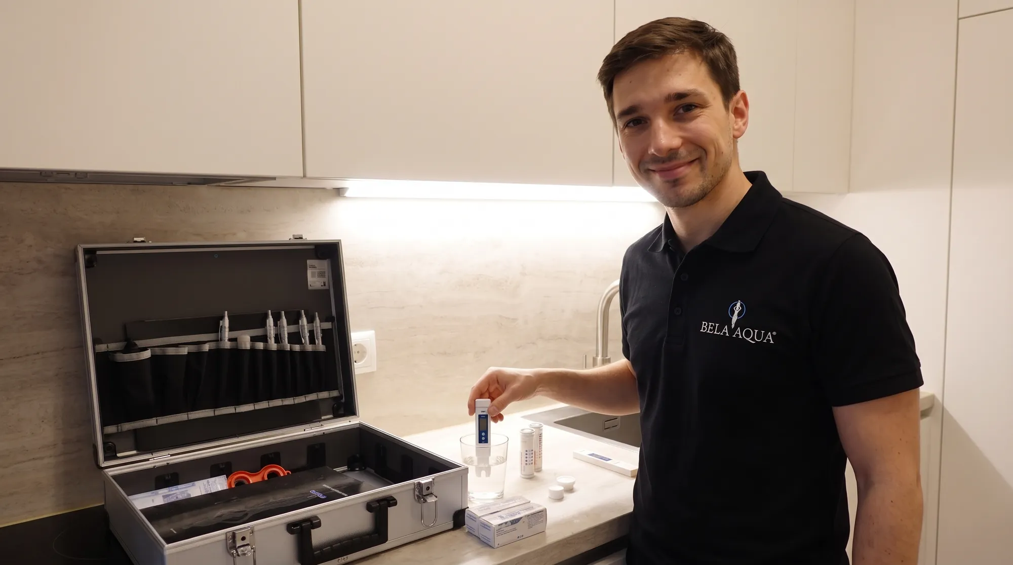 Christoph Speiser beim Wassertest in einer Küche: Messgerät im Wasserglas, daneben Teststreifen und der Testkoffer